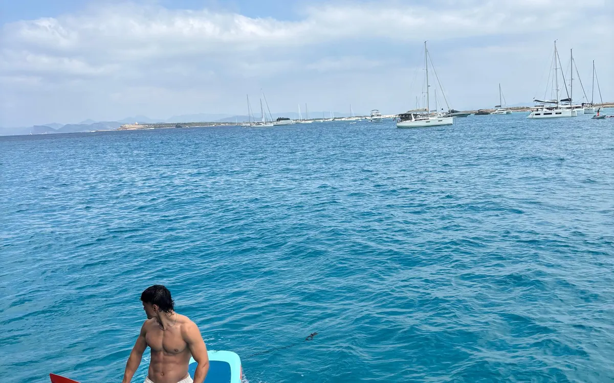 A guest sitting on the stern with a paddle board, sailing yachts moored across the bay behind