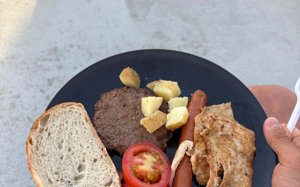 A breakfast plate with bread, a sausage, a burger, potatoes and tomato