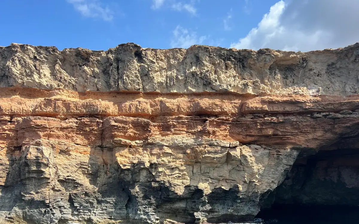 Layered sandy, red and grey cliffs above the sea, with a dark cave at the waterline