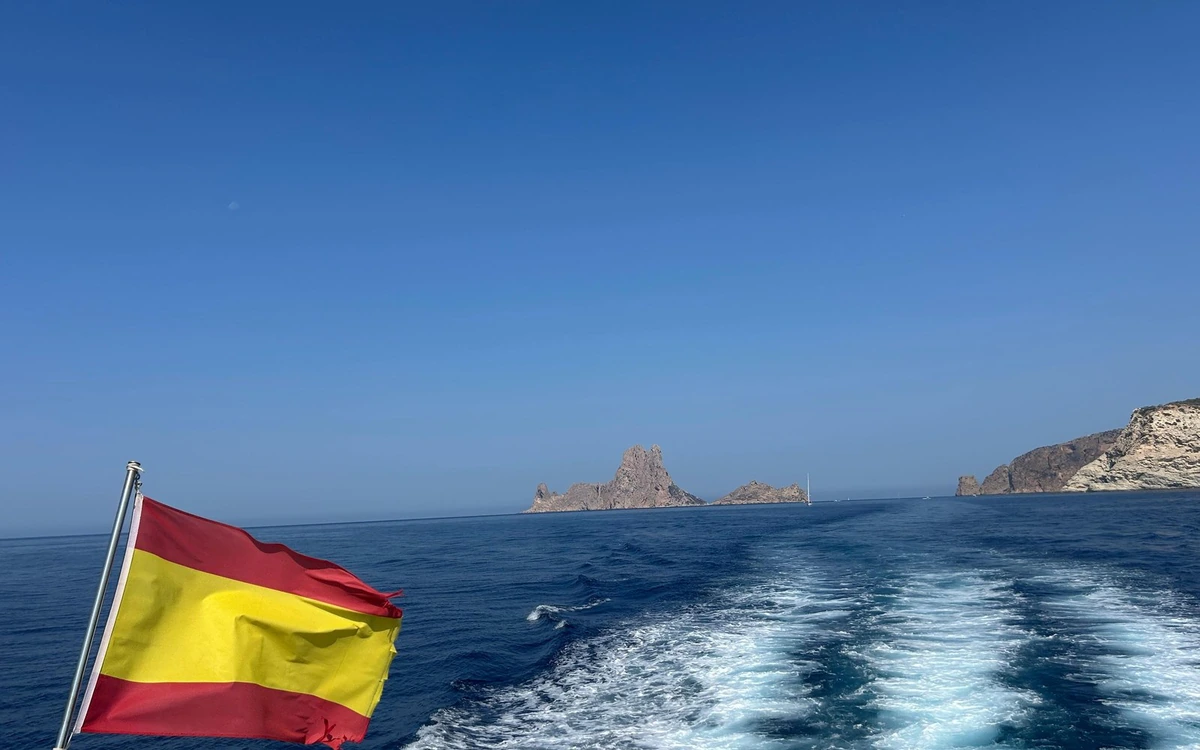 A Spanish flag flying over the white wake of a boat, a rocky islet on the horizon