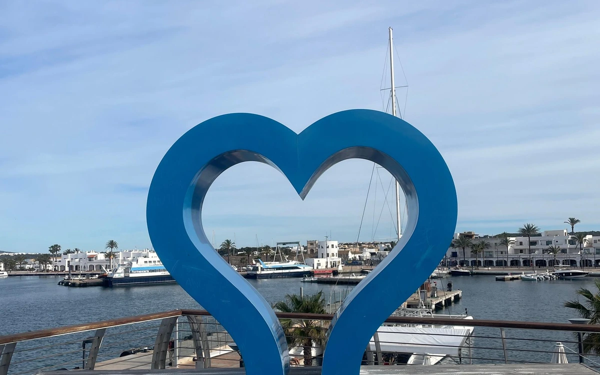 A large blue heart sculpture on a harbour front, ferries moored in the water behind it