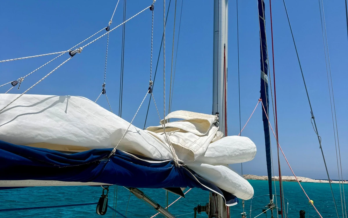 The furled white sail on the boom of a sailboat, turquoise water and a low coast behind
