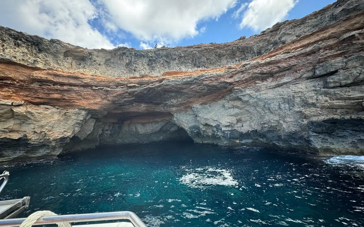 A sea cave under layered red and grey cliffs, seen from the rail of a boat over deep blue water