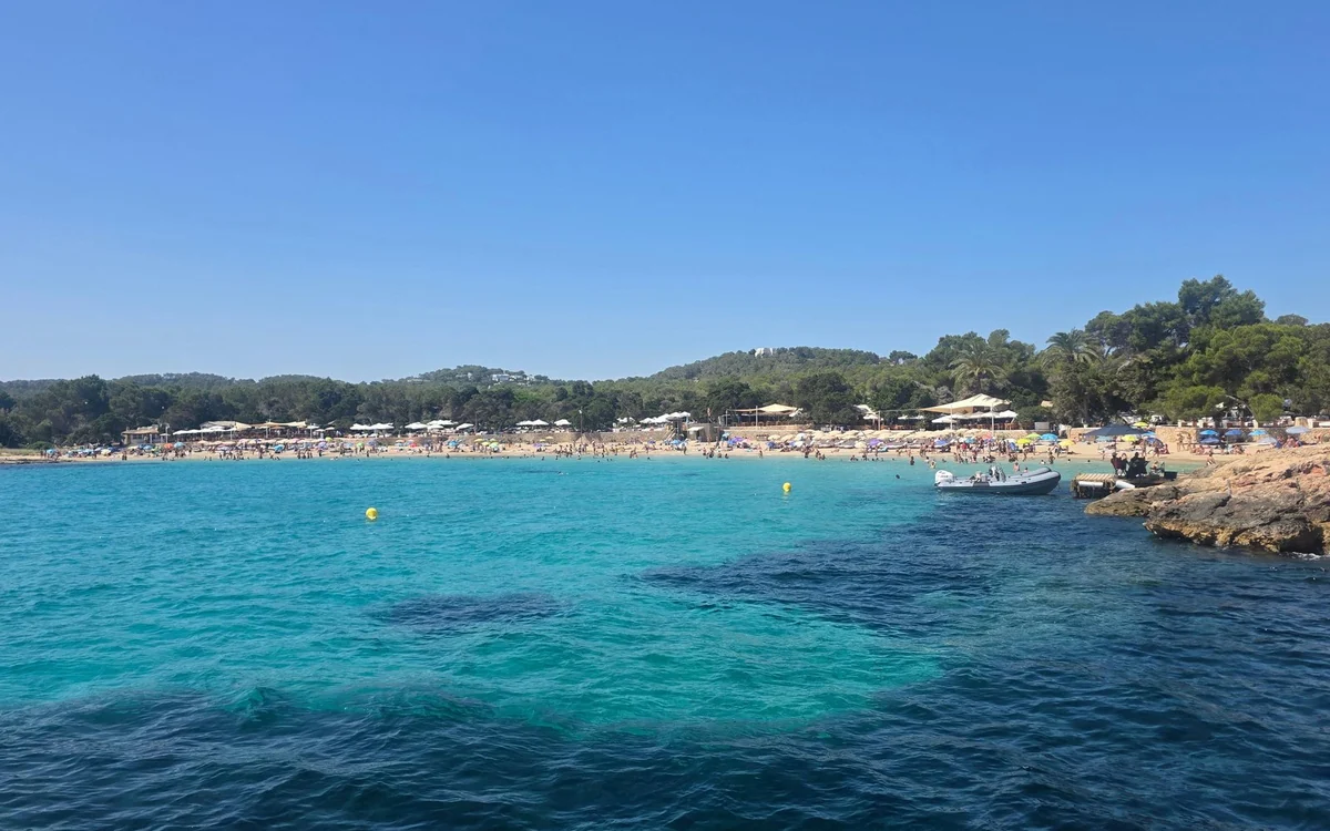 A sandy cove with sun umbrellas and pine-covered hills behind turquoise water, seen from a boat